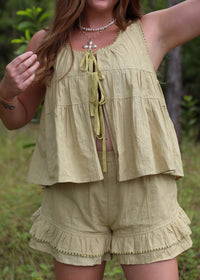A woman wearing a matching light olive green two-piece set consisting of a sleeveless ruffled crop top with tie-front detailing and matching ruffled shorts, photographed outdoors in a grassy, wooded setting.