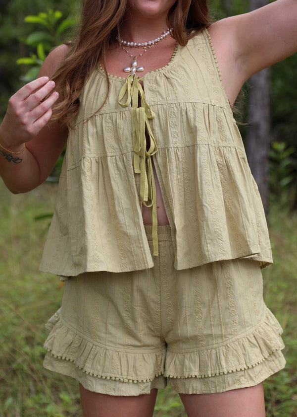 A woman wearing a matching light olive green two-piece set consisting of a sleeveless ruffled crop top with tie-front detailing and matching ruffled shorts, photographed outdoors in a grassy, wooded setting.