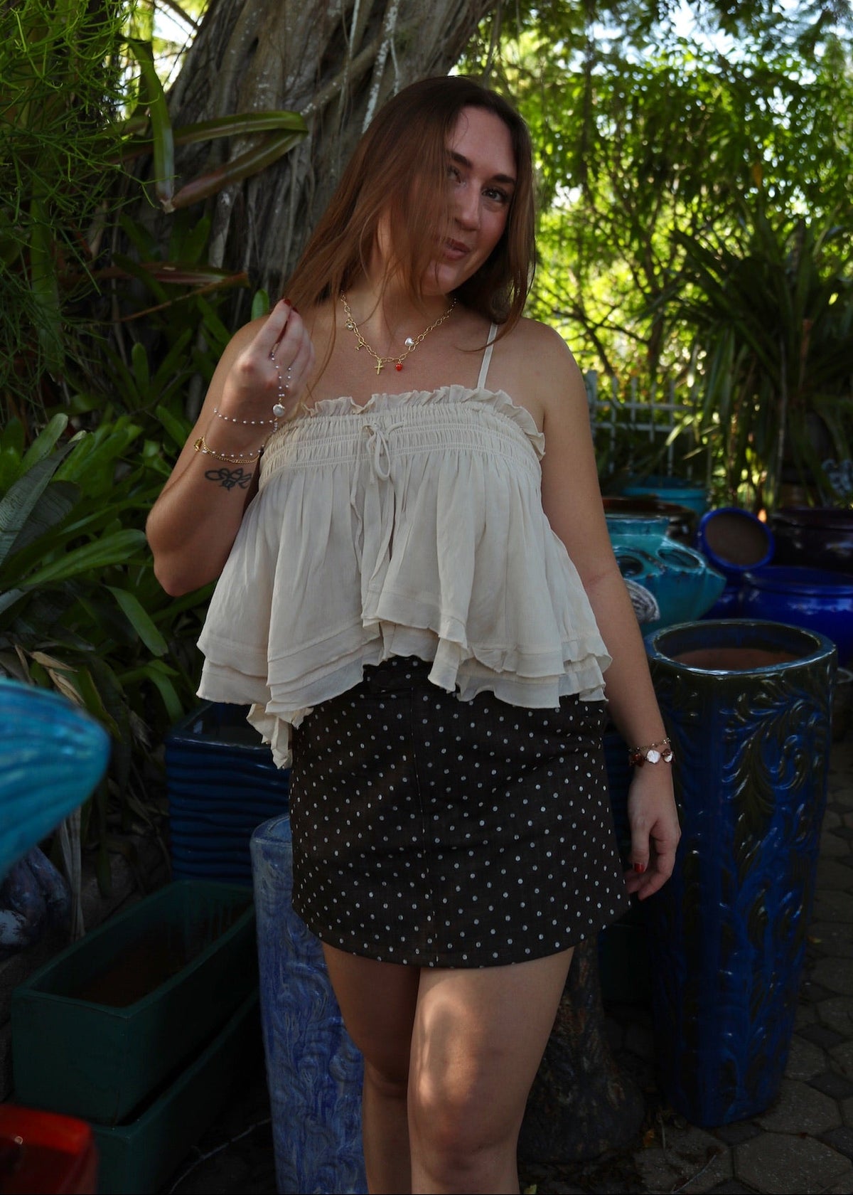 A woman posing outdoors in a garden setting, wearing a cream ruffled spaghetti-strap top and a black mini skirt with white polka dots.