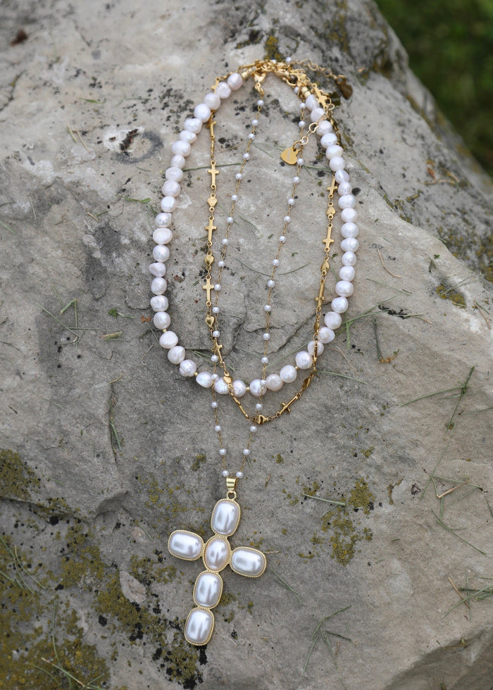 A double-strand pearl and gold necklace with a pearl cross pendant, displayed on a textured gray stone outdoors.