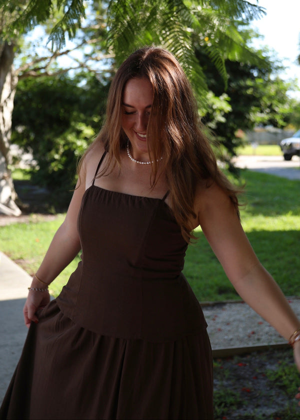 A woman wearing a dark brown spaghetti-strap midi dress, standing outdoors in a park-like setting with trees and grass in the background.
