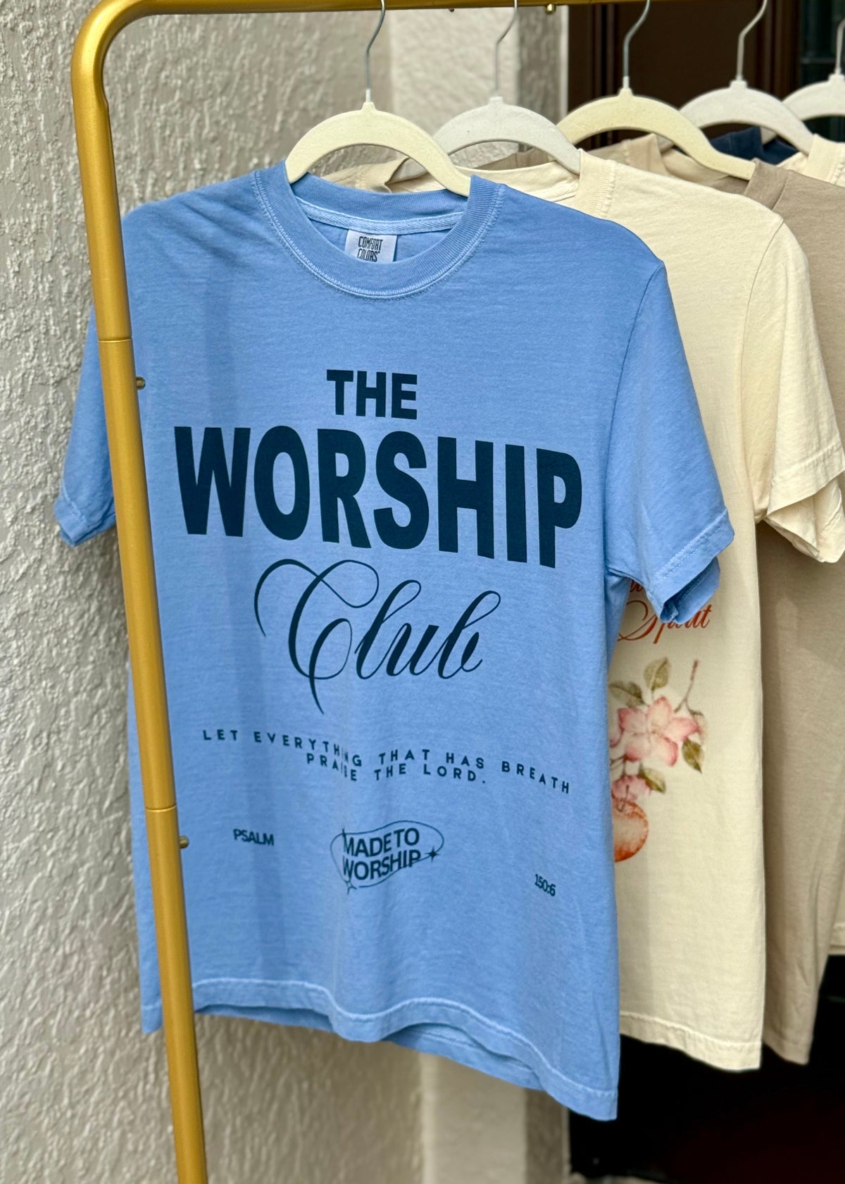 Blue t-shirt with 'The Worship Club' text on a rack with other shirts.