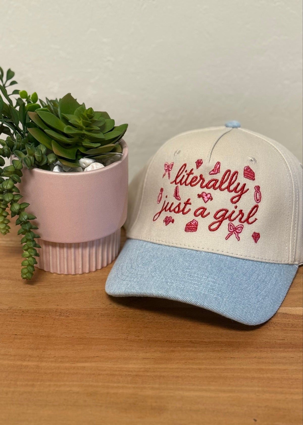 A baseball cap with a cream-colored crown and a light blue visor, featuring the embroidered text 'literally just a girl' in red script with small red hearts and bows. The cap is styled on a wooden surface next to a pink succulent pot against a textured off-white wall.