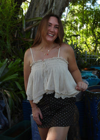 A woman smiling outdoors in front of a large tree, wearing a cream ruffled spaghetti-strap top and a black mini skirt with white polka dots.