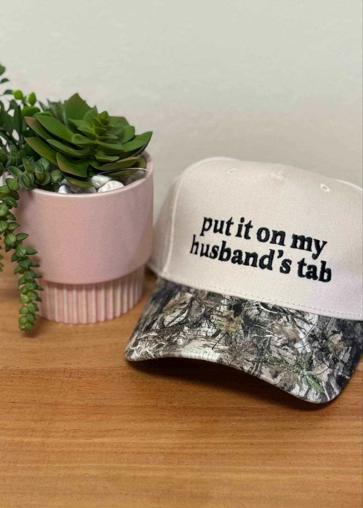 A white baseball cap with black text reading 'put it on my husband's tab' sits on a wooden surface next to a pink planter with a green succulent.