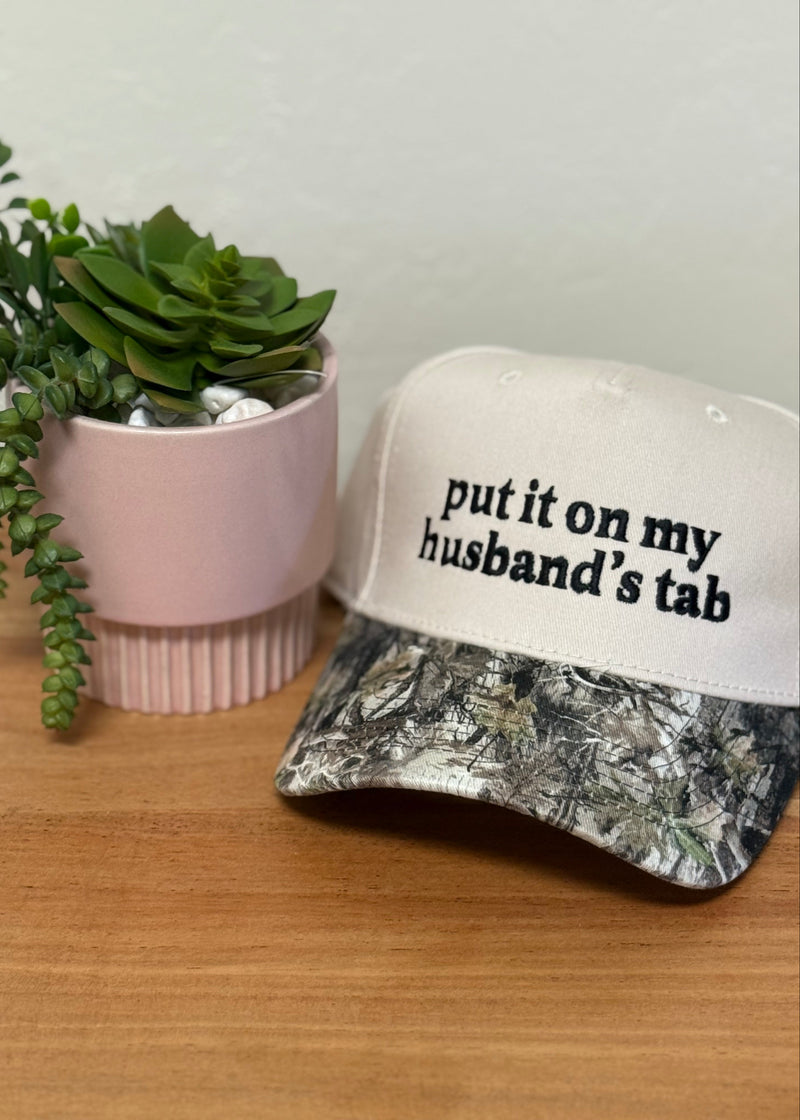 A white baseball cap with black text reading 'put it on my husband's tab' sits on a wooden surface next to a pink planter with a green succulent.