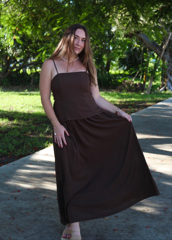 A woman wearing a dark brown sleeveless maxi dress stands on a paved path outdoors under leafy trees, holding out the side of the dress to show its flow.