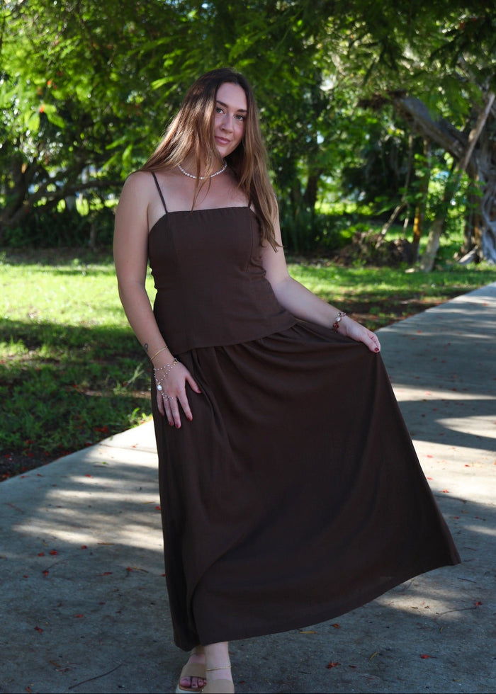 A woman wearing a dark brown sleeveless maxi dress stands on a paved path outdoors under leafy trees, holding out the side of the dress to show its flow.