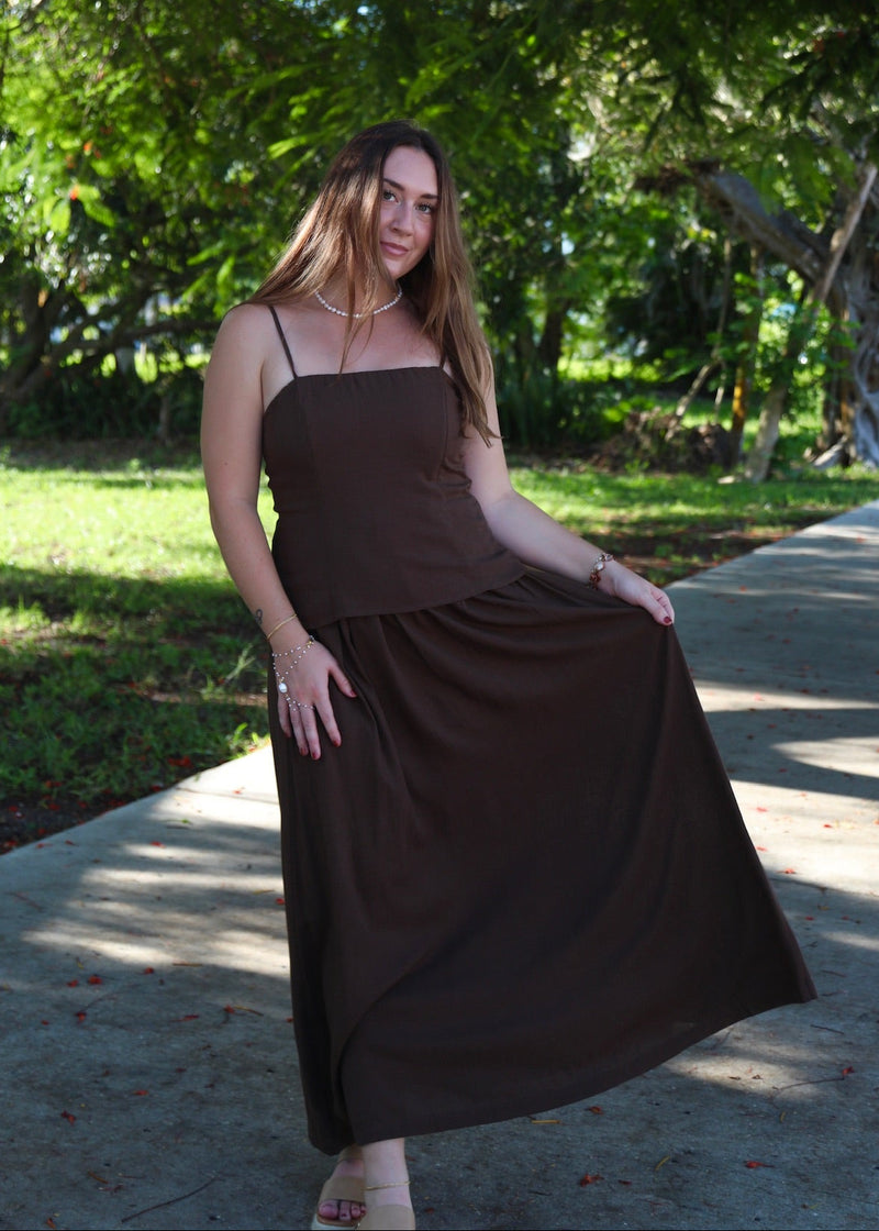 A woman wearing a dark brown sleeveless maxi dress stands on a paved path outdoors under leafy trees, holding out the side of the dress to show its flow.