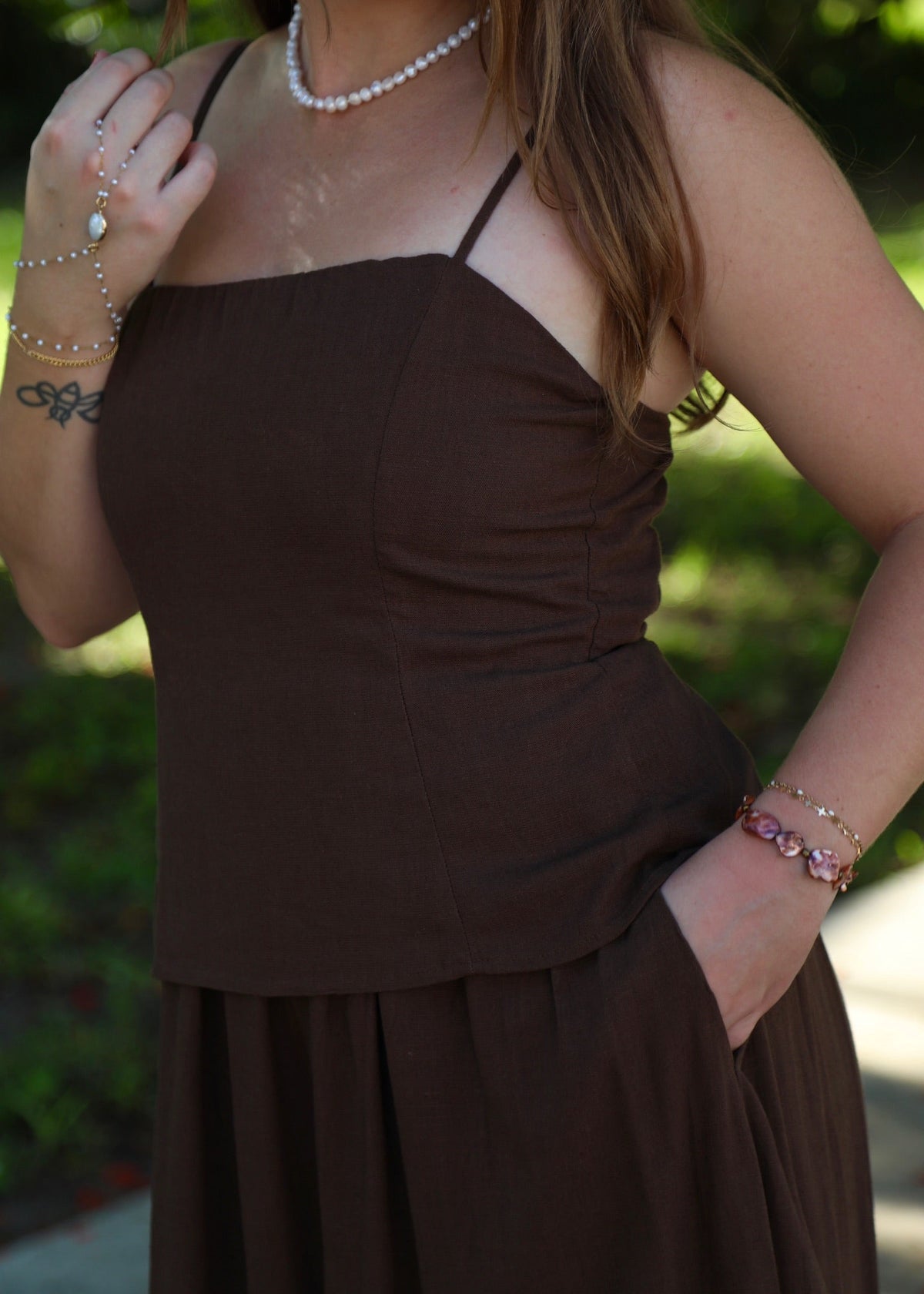 A woman wearing a dark brown spaghetti-strap dress with a ruched bodice and a flowing skirt, photographed outdoors with a blurred green background.