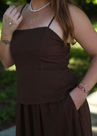 A woman wearing a dark brown spaghetti-strap dress with a ruched bodice and a flowing skirt, photographed outdoors with a blurred green background.