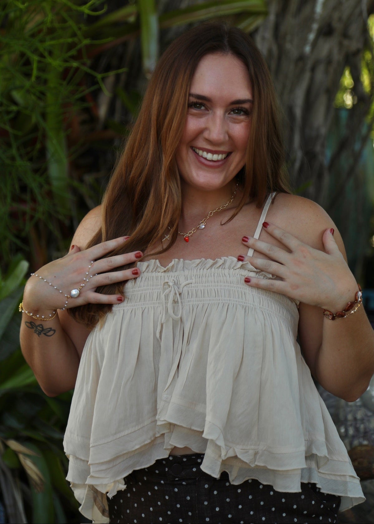A smiling woman with long brown hair poses outdoors wearing a cream-colored ruffled spaghetti-strap top and a black-and-white polka dot skirt, with green foliage and a tree trunk in the background.