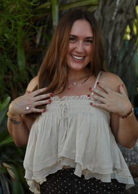 A smiling woman with long brown hair poses outdoors wearing a cream-colored ruffled spaghetti-strap top and a black-and-white polka dot skirt, with green foliage and a tree trunk in the background.