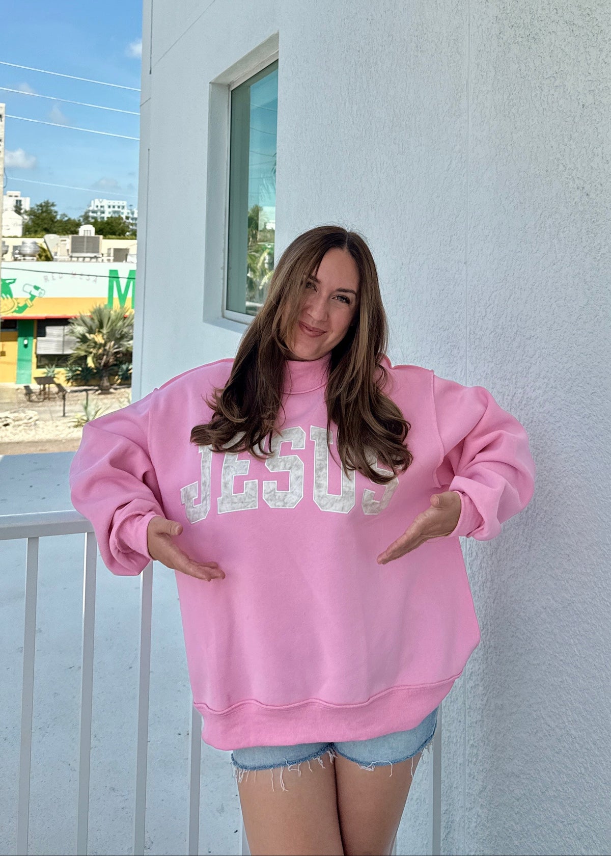 Person wearing a pink sweatshirt with 'JESUS' printed on it, standing on a balcony.