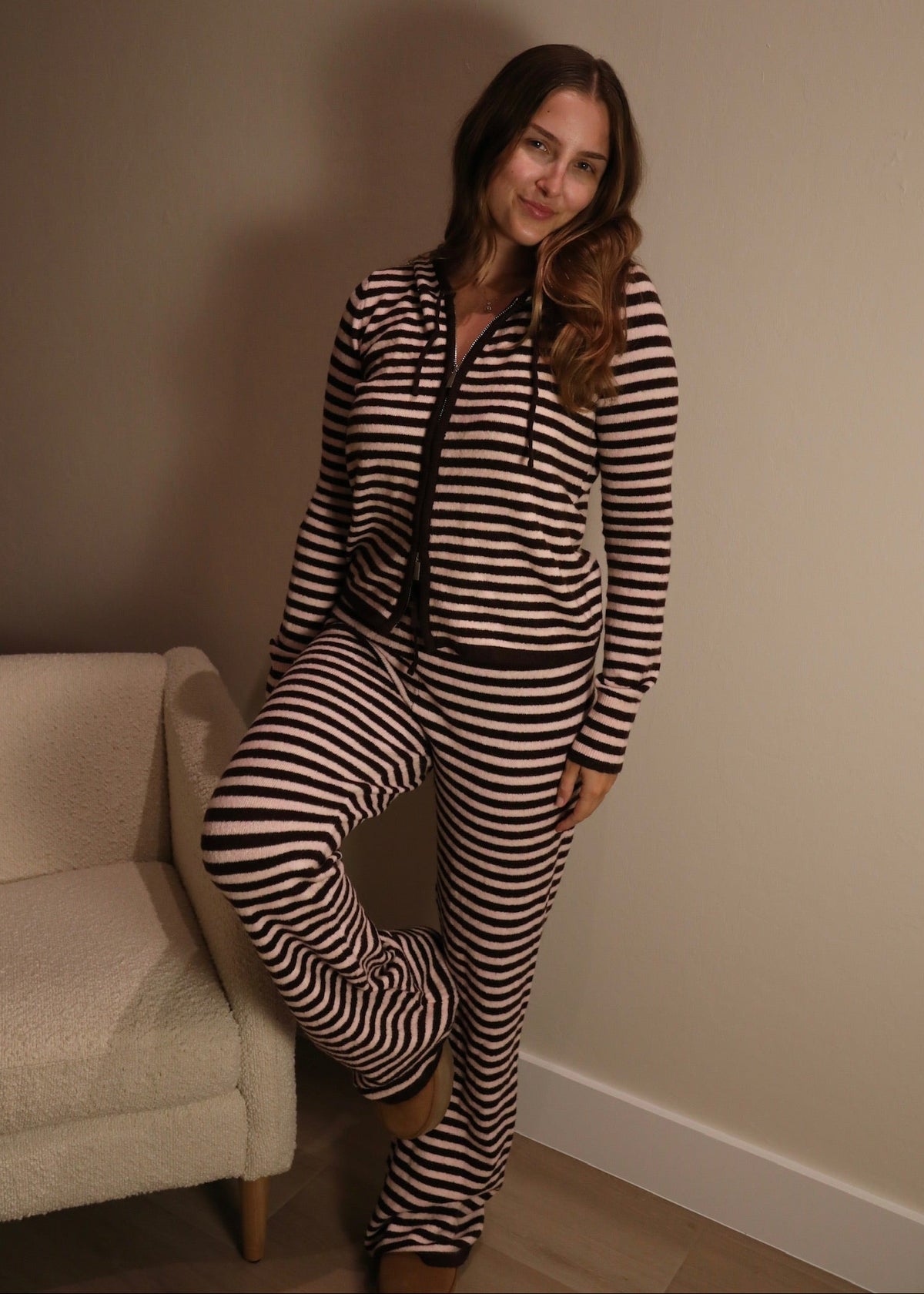 A woman wearing a matching two-piece loungewear set with black and beige horizontal stripes, consisting of a zip-up hoodie and sweatpants, posing in a room corner next to a cream-colored armchair.