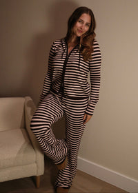 A woman wearing a matching two-piece loungewear set with black and beige horizontal stripes, consisting of a zip-up hoodie and sweatpants, posing in a room corner next to a cream-colored armchair.