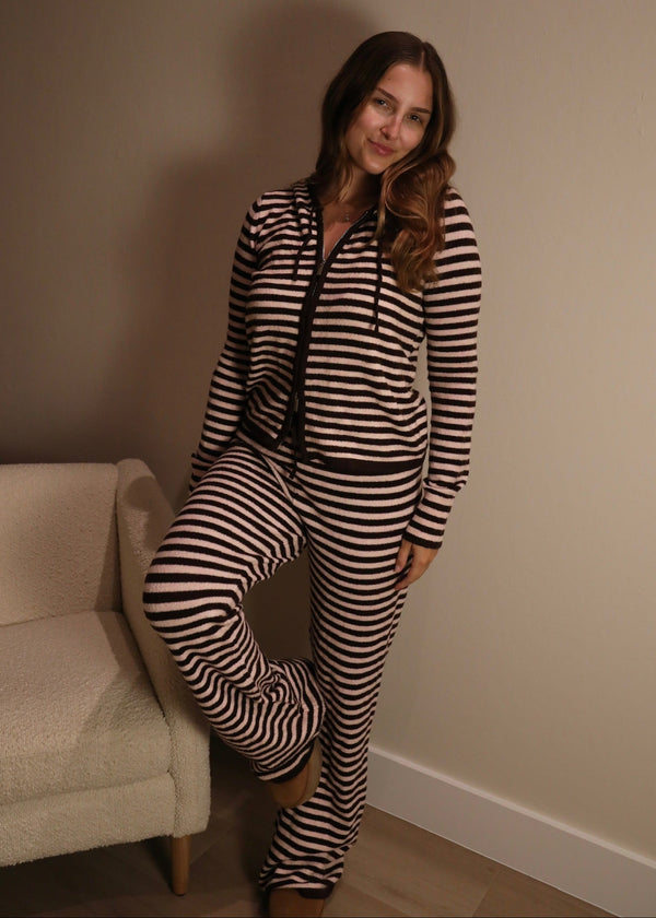 A woman wearing a matching two-piece loungewear set with black and beige horizontal stripes, consisting of a zip-up hoodie and sweatpants, posing in a room corner next to a cream-colored armchair.