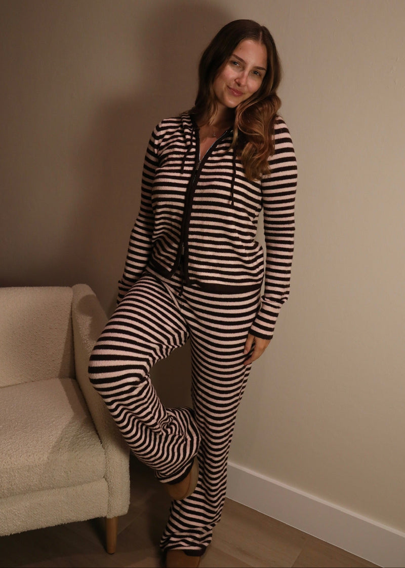 A woman wearing a matching two-piece loungewear set with black and beige horizontal stripes, consisting of a zip-up hoodie and sweatpants, posing in a room corner next to a cream-colored armchair.