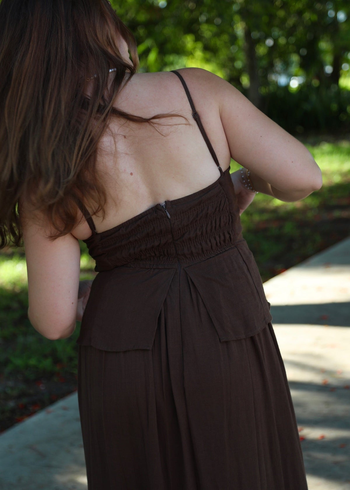 A woman wearing a dark brown spaghetti-strap maxi dress, photographed from behind in an outdoor park setting with green trees and a concrete path.