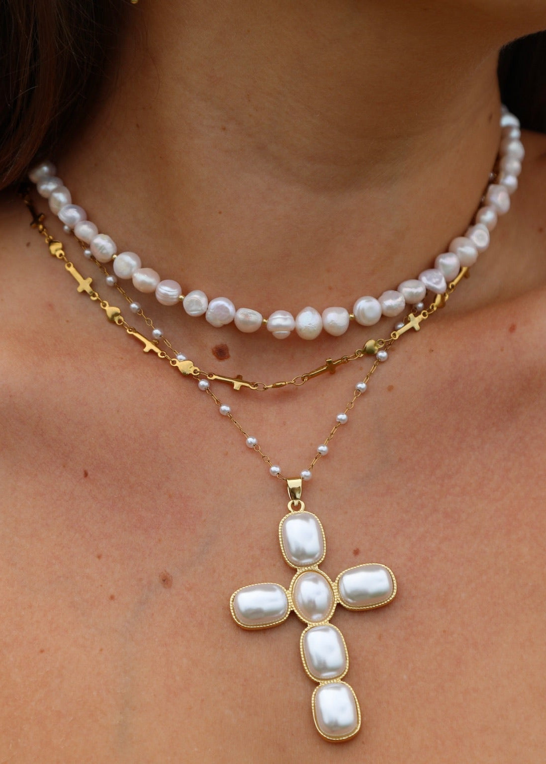 A close-up of a woman's neck and shoulders wearing a layered necklace set. The set features a top strand of white baroque pearls, a middle gold chain with small cross charms, and a lower chain with tiny pearls leading to a larger white baroque pearl cross pendant.