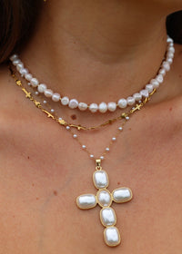 A close-up of a woman's neck and shoulders wearing a layered necklace set. The set features a top strand of white baroque pearls, a middle gold chain with small cross charms, and a lower chain with tiny pearls leading to a larger white baroque pearl cross pendant.