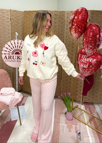 Woman in a white sweater with heart designs and pink pants standing next to a display of heart-shaped balloons in a room with a pink chair and decorative items.