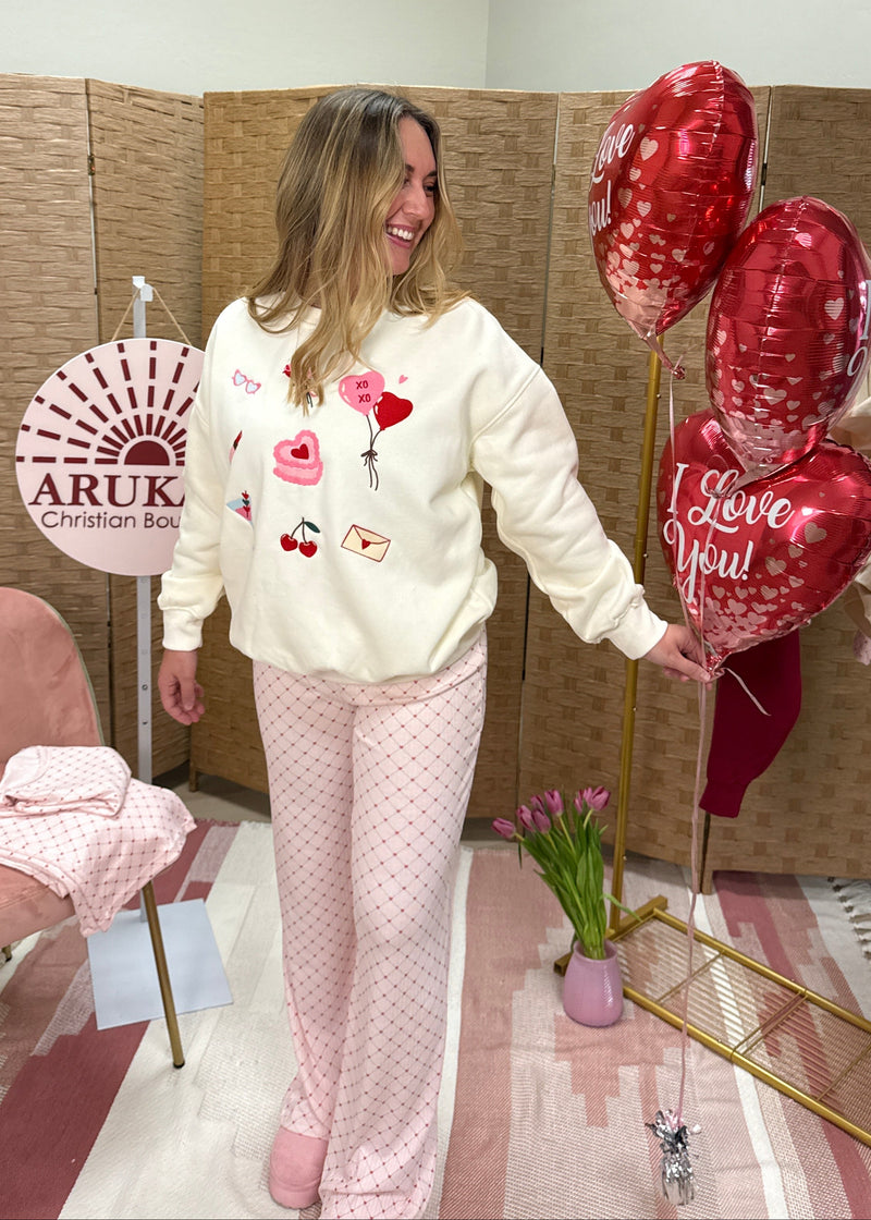 Woman in a white sweater with heart designs and pink pants standing next to a display of heart-shaped balloons in a room with a pink chair and decorative items.