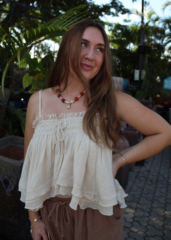 A woman posing outdoors in a cream ruffled spaghetti-strap top and brown pants, wearing a red beaded necklace. The background features lush green foliage and a paved walkway.