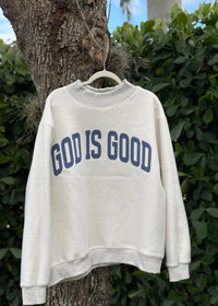 White sweatshirt with 'GOD IS GOOD' text hanging on a tree branch outdoors.