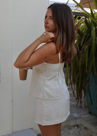 A woman wearing a light beige linen-look two-piece set consisting of a sleeveless top and matching mini skirt, standing outdoors next to a white wall and green plants.
