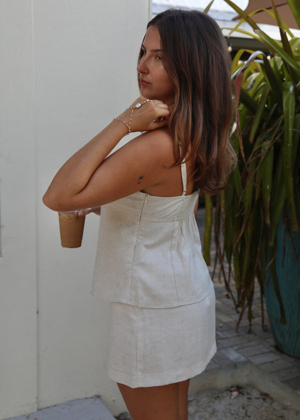 A woman wearing a light beige linen-look two-piece set consisting of a sleeveless top and matching mini skirt, standing outdoors next to a white wall and green plants.