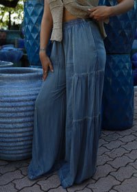 A person wearing a beige cropped knit top and high-waisted, wide-leg blue denim maxi pants, standing outdoors next to large blue ceramic pots on a paved surface.
