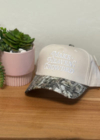 A beige trucker-style cap with a camouflage-patterned brim and the phrase 'MAKE HEAVEN CROWDED' embroidered on the front, displayed next to a small potted succulent on a wooden surface.