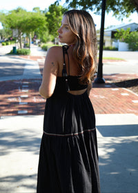 A woman wearing a black sleeveless maxi dress with a cutout back, photographed outdoors on a sunny sidewalk with trees and a street in the background.