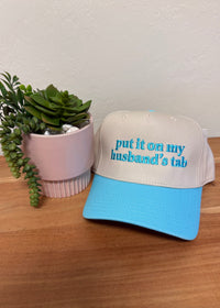 A light blue and white trucker hat with the phrase 'put it on my husband's tab' embroidered in blue on the front, displayed on a wooden surface next to a small potted succulent.