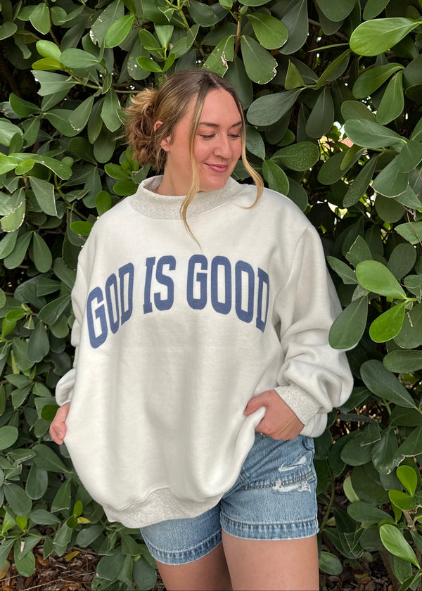 Person wearing a 'God is Good' hoodie standing among green bushes