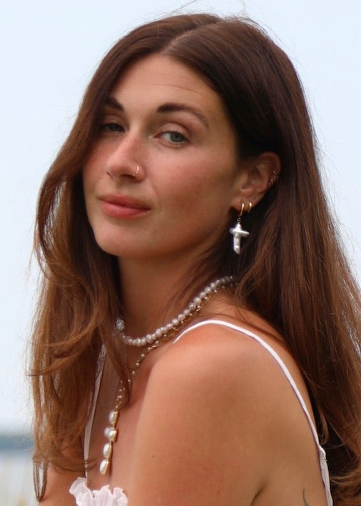 A woman with long brown hair stands outdoors by a wooden fence overlooking water, wearing a white ruffled sleeveless top and dark gray cargo pants.