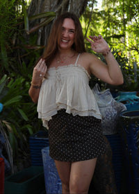 A woman posing outdoors in front of a large tree, wearing a cream ruffled spaghetti-strap top and a black polka-dot mini skirt.