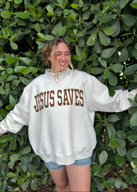 Person wearing a grey sweatshirt with 'Jesus Saves' text,standing in front of green leaves