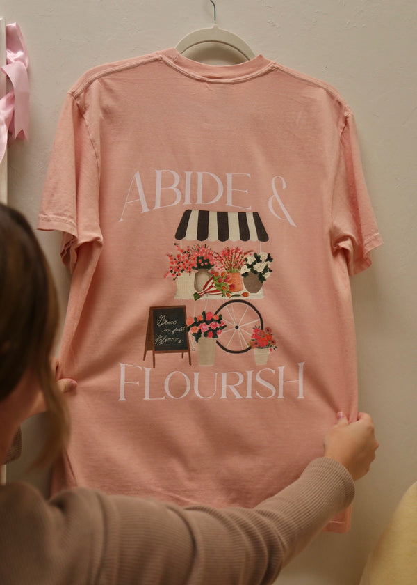 Pink t-shirt with 'Abide & Flourish' text and design on a hanger