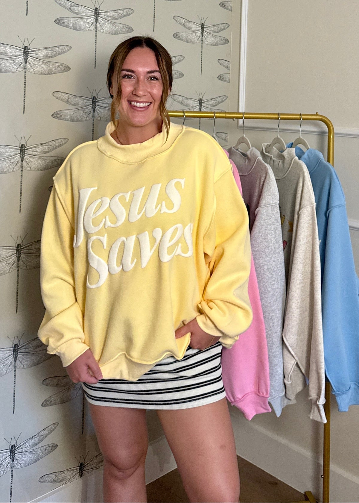 Person wearing a yellow 'Jesus Saves' sweatshirt in a room with dragonfly wallpaper.