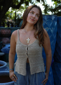 A woman wearing a beige knit sleeveless top with a button-front detail and a blue skirt, standing outdoors in front of blue ceramic pots.