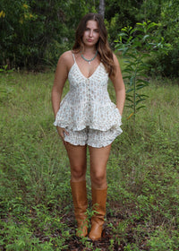 A woman stands in a grassy wooded outdoor setting wearing a white floral camisole top and matching ruffled shorts, paired with tan knee-high boots.